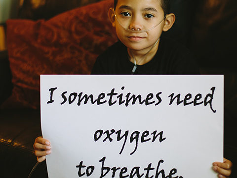 A boy with oxygen supply tube holding a sign saying "I sometimes need oxygen to breathe."